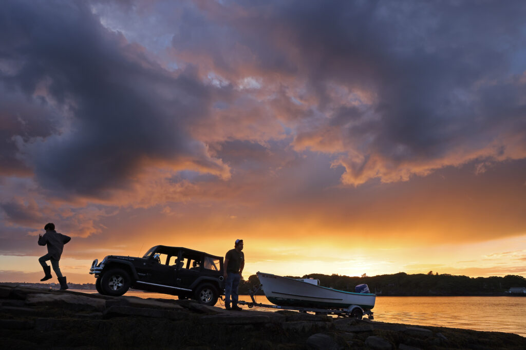 In the background a summer sunset, in the foreground a boy runs up a boat ramp while a man hauls a boat out of the water